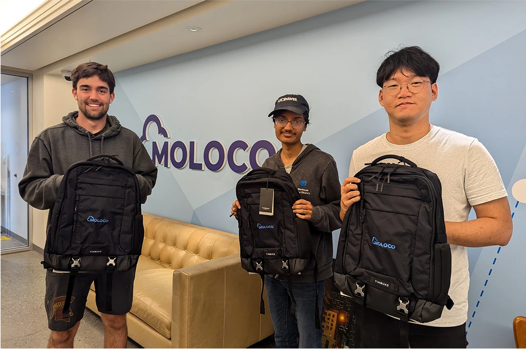 Three Moloco interns in a modern office setting, each holding a black backpack with a 'Moloco' logo, stand in front of a wall with the company name.
